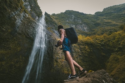 一名身穿蓝色短裤的男子站在岩层上，手持数码单反相机站在瀑布前